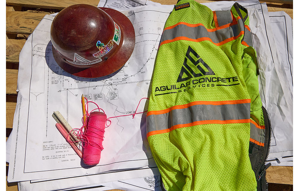A red hard hat, neon safety vest with the Aguilar Concrete Services logo, pink string, and markers rest on top of construction blueprints laid out on a wooden surface at a job site.